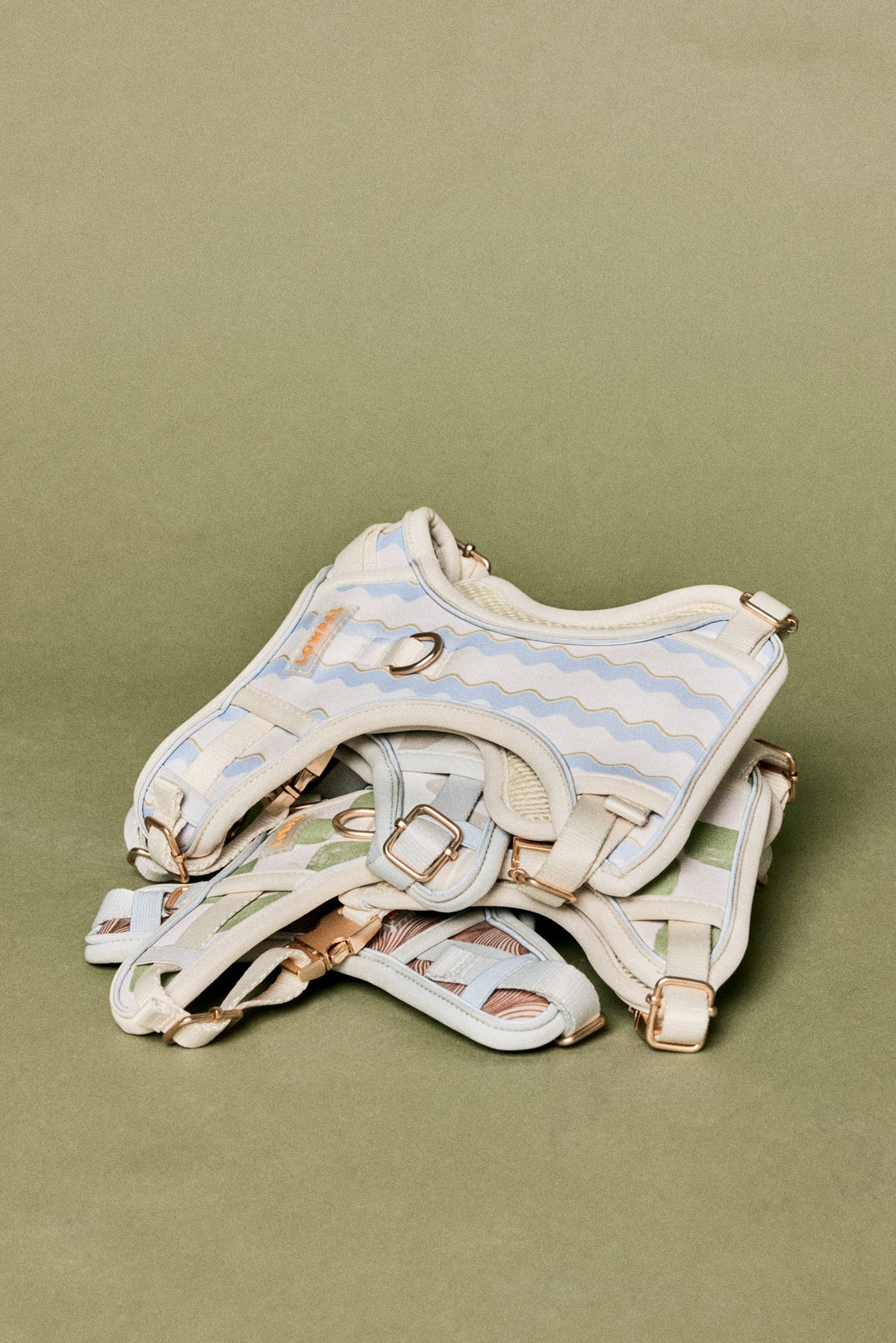 A dog harness with a cream and light blue wavy stripe pattern, accented with rose gold hardware, displayed on a solid olive green background.